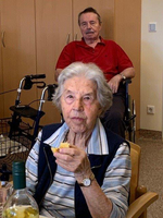 Eine ältere Frau isst, während ein Mann im Hintergrund sitzt.
