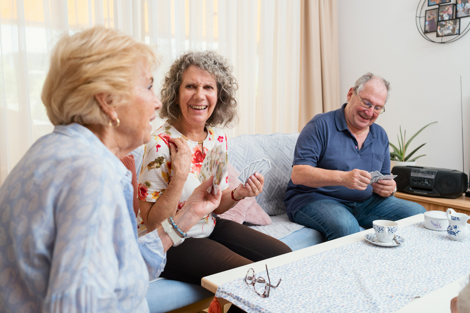 Drei Senioren der Wohngemeinschaft sitzen zusammen auf einer Couch und spielen ein Kartenspiel, während sie eine Tasse Kaffee genießen. Alle wirken entspannt und amüsiert
