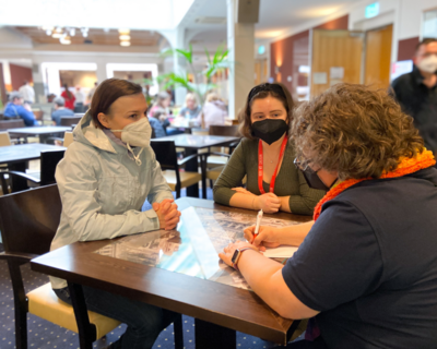 drei Personen an einem Tisch, alle tragen FFP2-Maske, zwei sind von vorne bzw. seitlich zu sehen, dritte Person nur zum Teil und von hinten ersichtlich, notiert etwas auf einen Zettel, im Hintergrund Raum mit weiteren Personen und Tischen, Pflanzen 