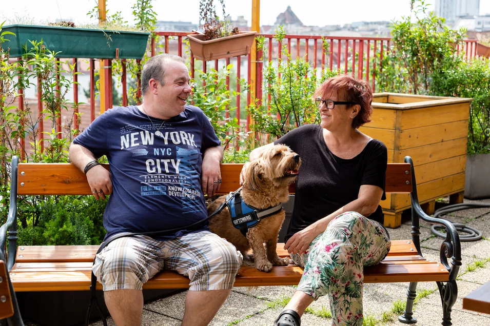 Eine Bewohnerin und ein Bewohner sitzen mit Hund auf Bank in Gemeinschaftsgarten