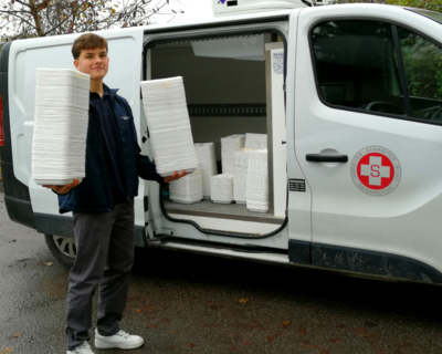 Zivildiener in dunkler Hose und mit blauer Fleecejacke mit Samariterbund Wien Logo vor offenem Kastenwagen, hält viele Boxen von Essen auf Rädern in beiden Händen hochgestapelt, weitere Boxen im Auto, auf Auto Samariterbund Logo auf Türe