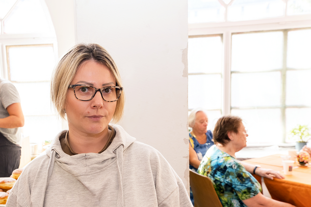 „Im Vordergrund steht eine Frau, die für die Kamera posiert. Im Hintergrund sind mehrere Personen zu sehen, von denen einige am Tisch sitzen, während andere ehrenamtlich bei der Essensausgabe dabei sind. Samariterbund-Suppentopf