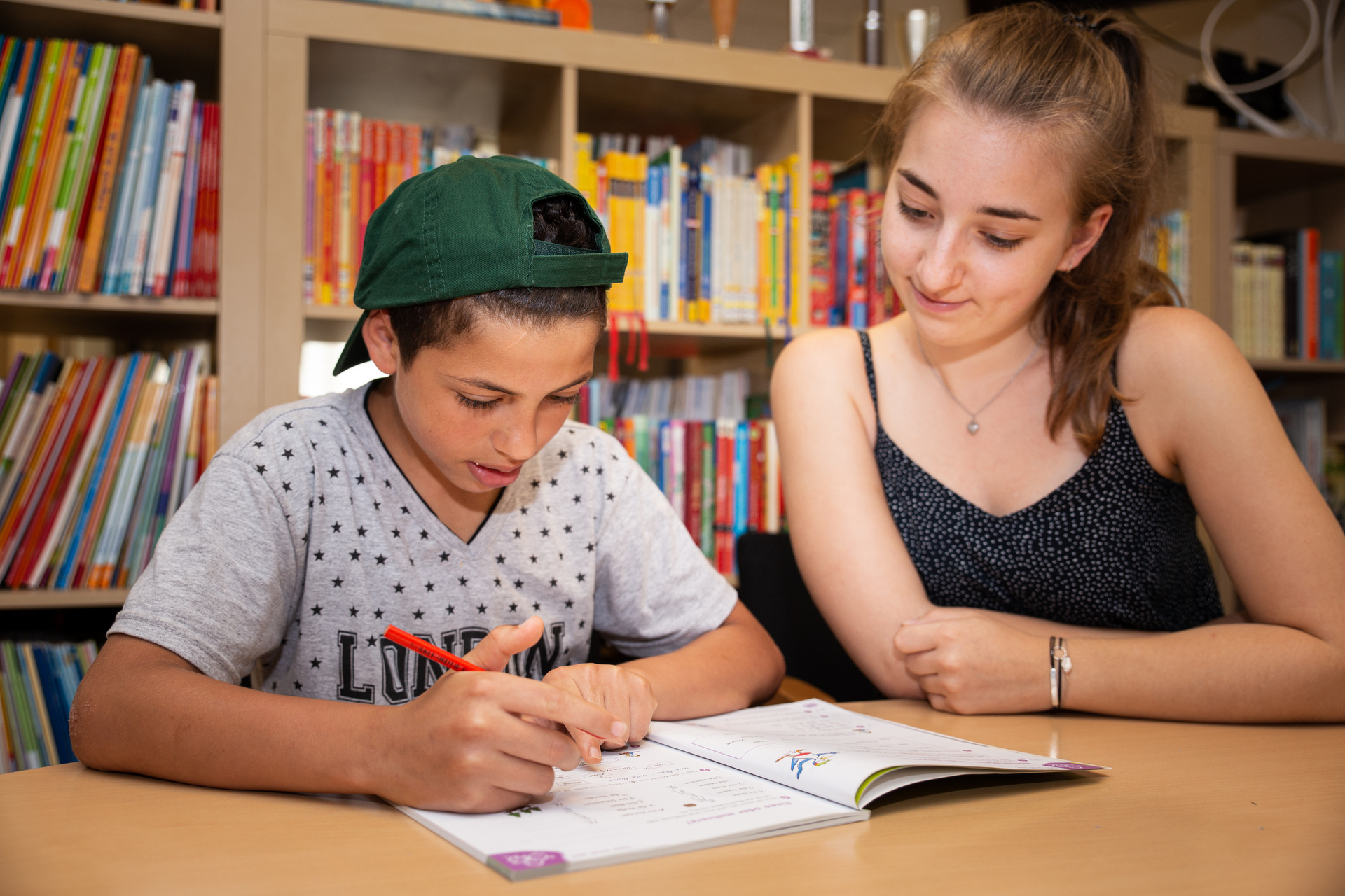 Mädchen und Junge sitzen an Schreibtisch vor Bücherregal und lernen gemeinsam