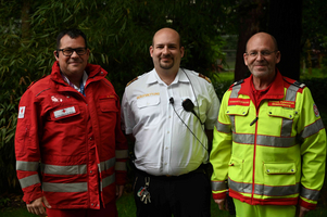Drei Männer stehen lächelnd in einem Garten. Der linke Mann trägt eine rote Jacke mit grauen Streifen, der mittlere ist in weißem Hemd mit einem Funkgerät und der rechte in leuchtend gelber Jacke mit reflektierenden Streifen. Hintergrund mit grünen Pflanzen.