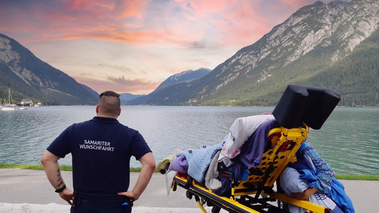 Wunschfahrt-Mitarbeiter mit Person auf Trage vor See und Bergen im Hintergrund