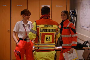 Drei Personen in einem Besprechungsraum des Samariters. Zwei Frauen in Rettungsdienstkleidung lächeln, während ein Mann mit einer Jacke des Einsatzleiters in der Mitte steht. Im Hintergrund sind Schreibtische und organisatorische Materialien zu sehen.