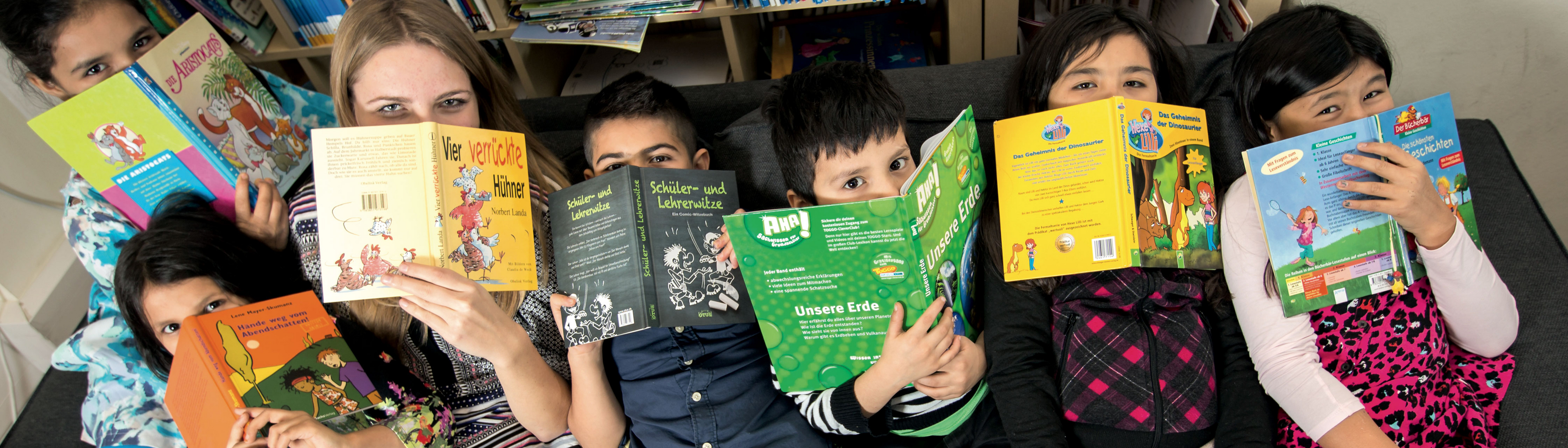 LernLEO - Sechs Kinder und eine Betreuerin liegen am Sofa und verstecken ihre Gesichter unterhalb der Nase mit Büchern
