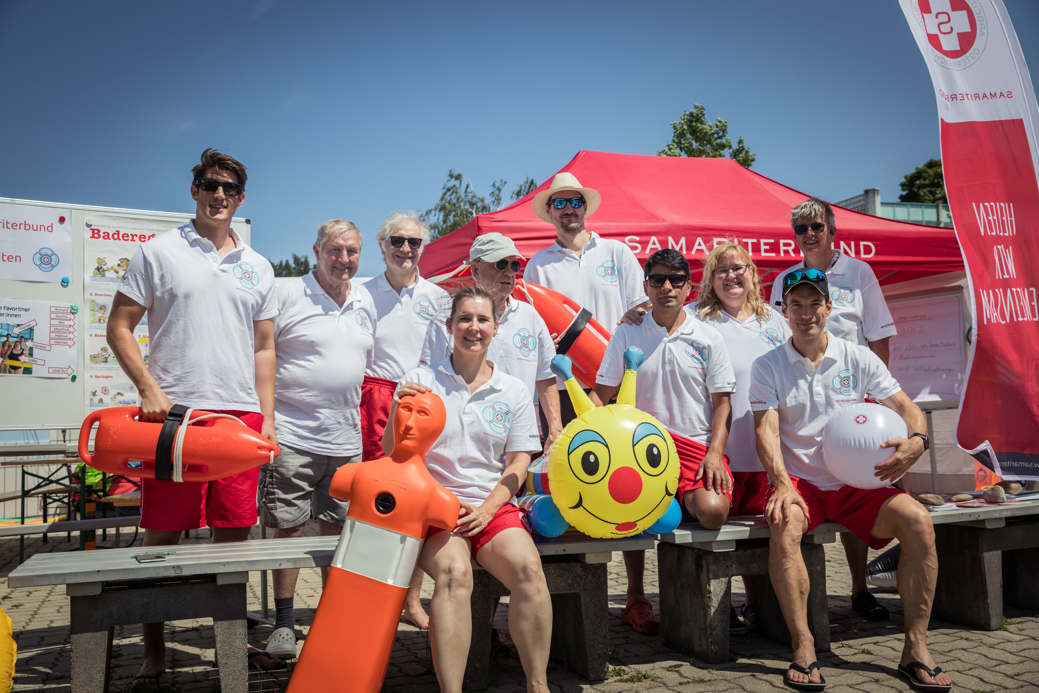 Gruppenbild Mitarbeiter:innen der Wasserrettung mit weißen T-Shirts, sitzen bzw. stehen als Gruppe beisammen, daneben Wasserrettungsgeräte, im Hintergrund rotes ZElt und Beachflag